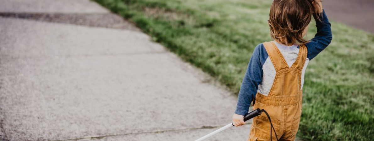 Vision impaired child in yellow dungarees walking along the pavement with a cane