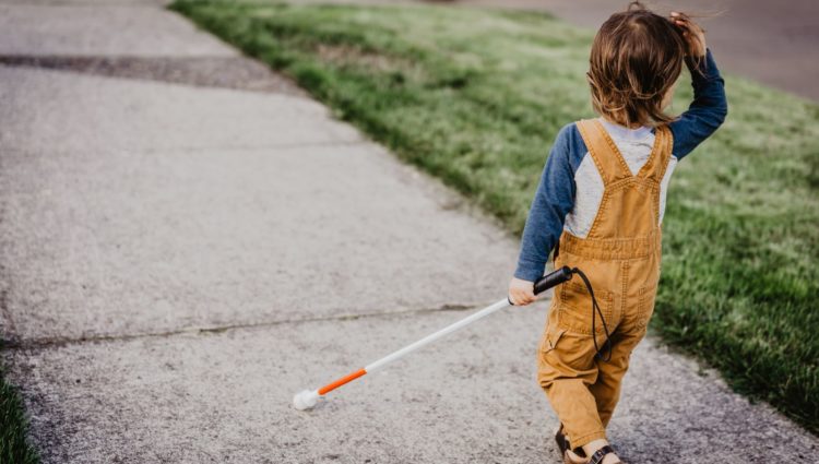Vision impaired child in yellow dungarees walking along the pavement with a cane