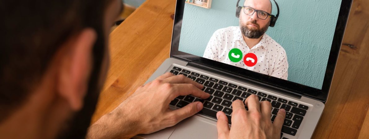 Over the shoulder shot of a person in a virtual meeting on a laptop with a man wearing headphones.