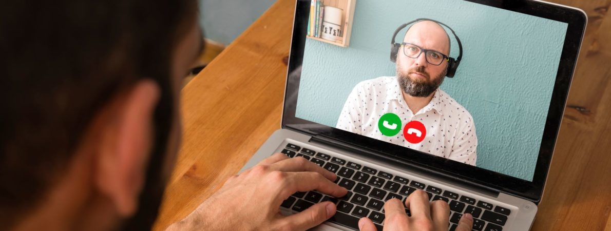 Over the shoulder shot of a person in a virtual meeting on a laptop with a man wearing headphones.