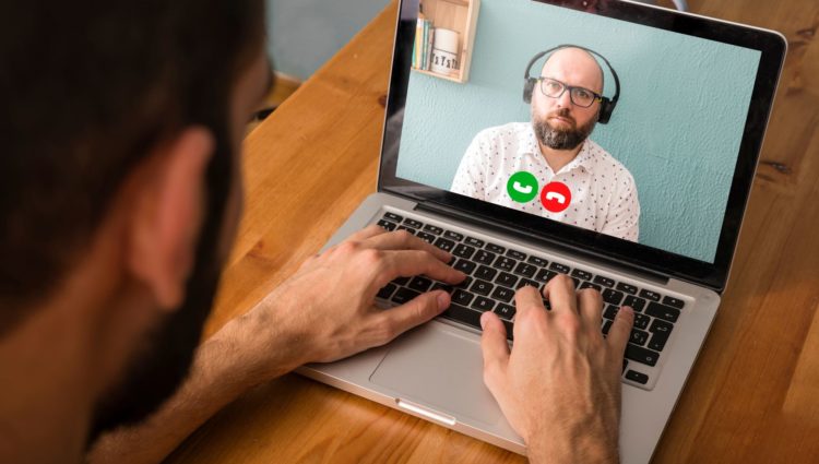 Over the shoulder shot of a person in a virtual meeting on a laptop with a man wearing headphones.