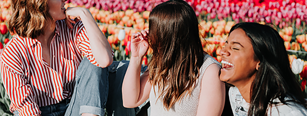 Three young women sat on a bench, one woman is laughing. There is a field of flowers in the background.