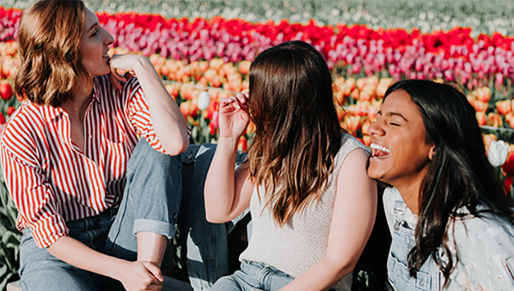 Three young women sat on a bench, one woman is lauging. There is a field of flowers in the background