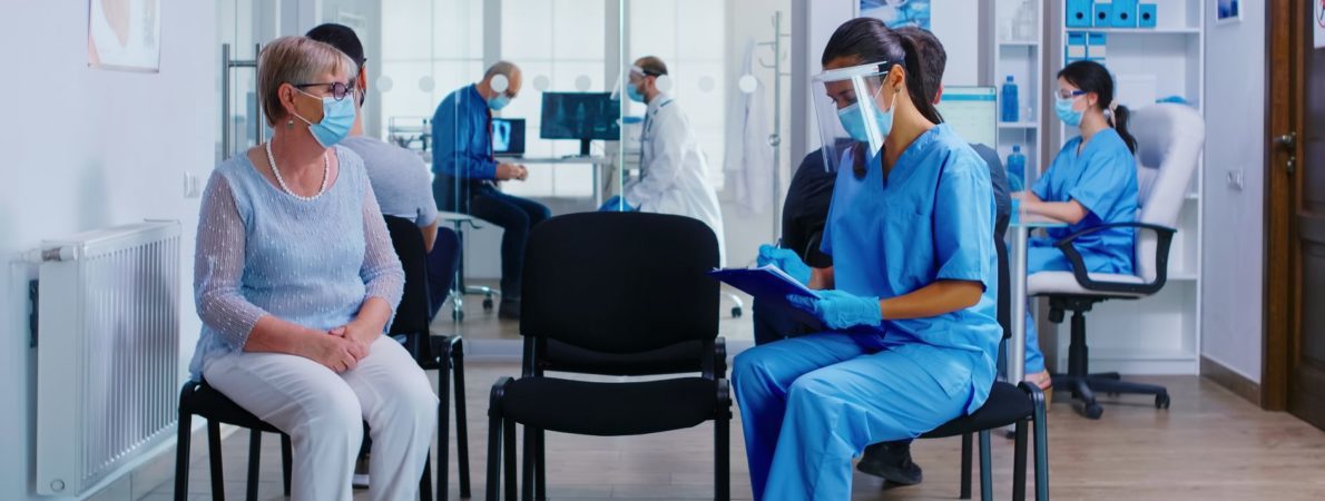 Hospital waiting room. Patient wearing a mask sits across from a nurse wearing a visor.