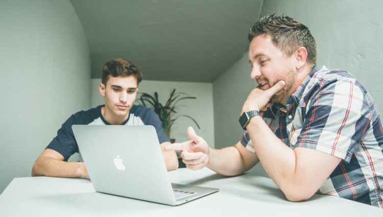 A teacher sat at a desk with a student, both looking at a laptop together and talking.