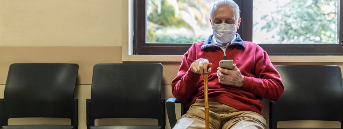 Elderly man wearing a face mask uses Aira app on smartphone in a hospital waiting room.