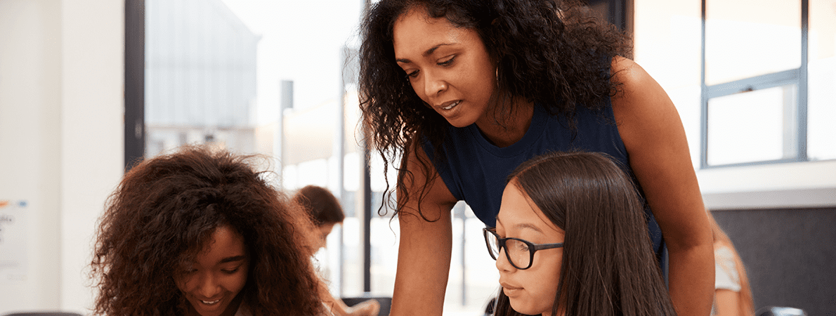 Teacher leaning over table talking to two young female students