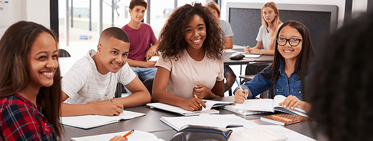 Group of diverse students in the classroom. They are facing the teacher. The teacher has their back to the camera.