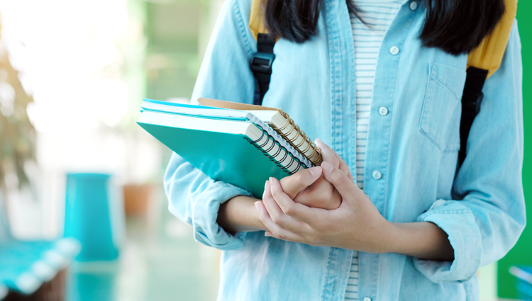 A close up of a female student holding a stack of notepads, walking down a university corridor