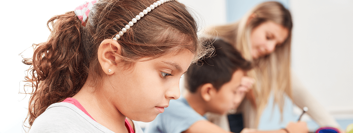 Girl in class looking at her notebook. There is a teacher in the background talking to a child.