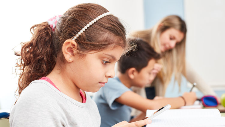 Girl in class looking at her notebook. There is a teacher in the background talking to a child.