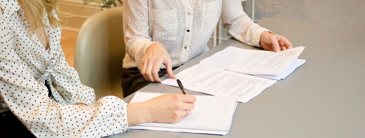 Two women sat at a desk writing on a stack of paper