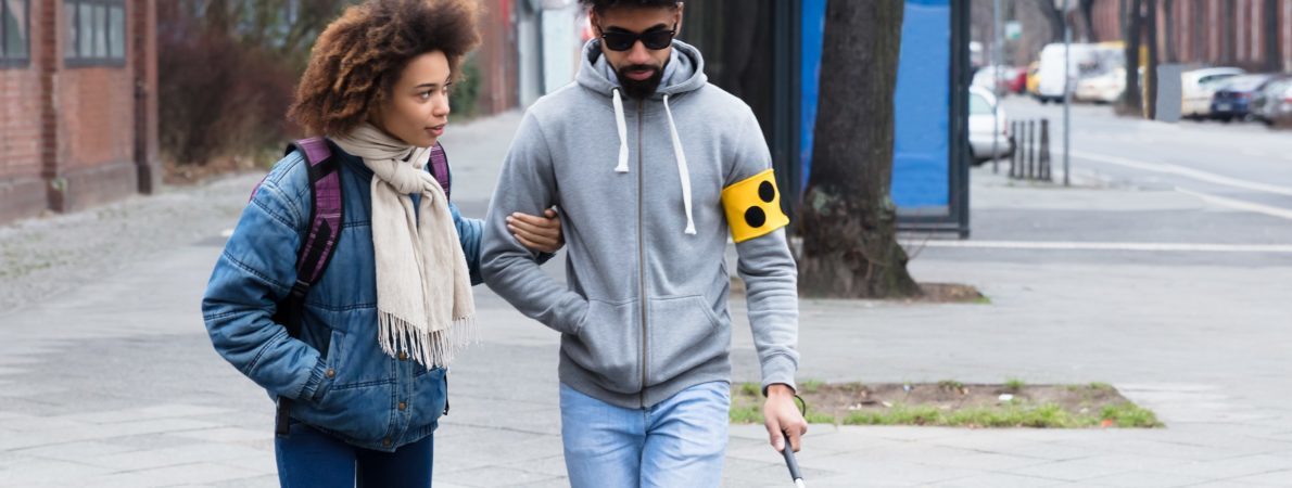 A young woman helping a young blind man. The man is holding a white cane.