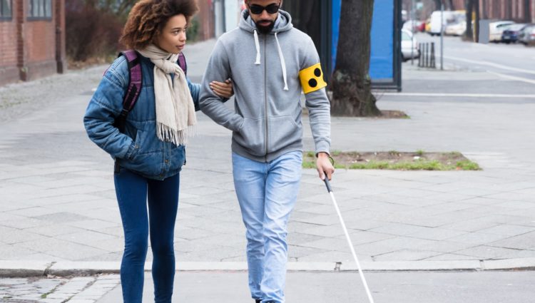A young woman helping a young blind man. The man is holding a white cane.