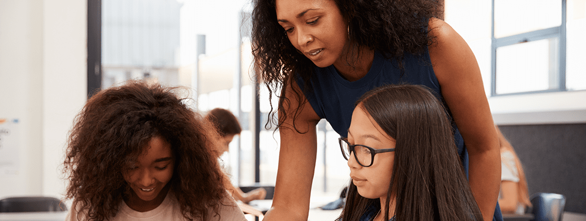 Teacher leaning over table talking to two young female students