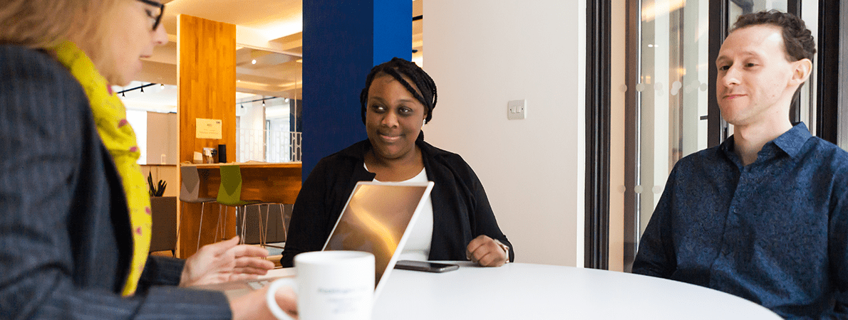 Woman talking to a blind young woman and man at a table. The woman is using a laptop.