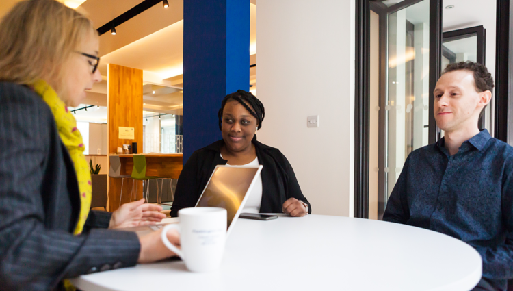 Woman talking to a blind young woman and man at a table. The woman is using a laptop.