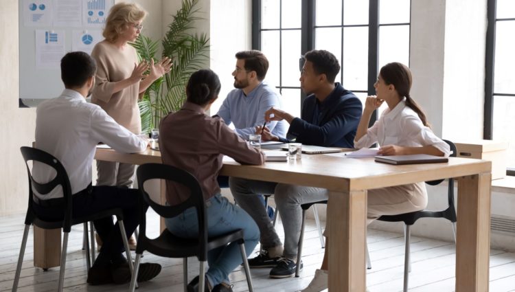 Group of people at a meeting room. They are gathered around the table listening to a woman giving a presentation in front of a board