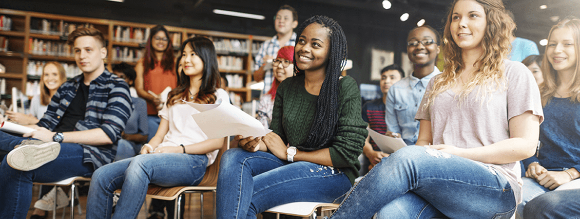 A large group of students sat in a crowded room, smiling and facing the front of the room
