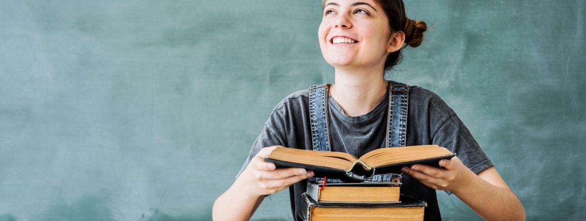 young student with a pile of books, holding a book, smiling and looking up