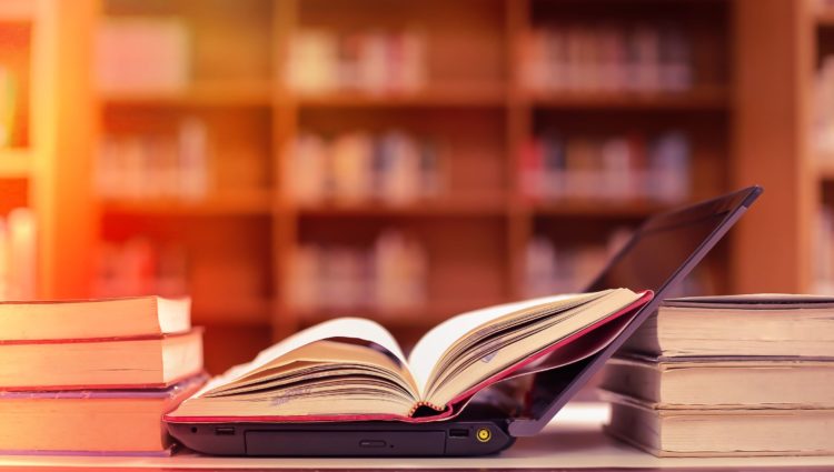 An open book on an open laptop, surrounded by books in a library