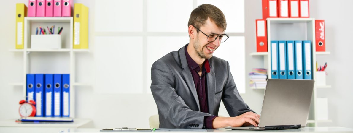 A young man sitting on a desk and working on his laptop, smiling, a pile of folders at the back