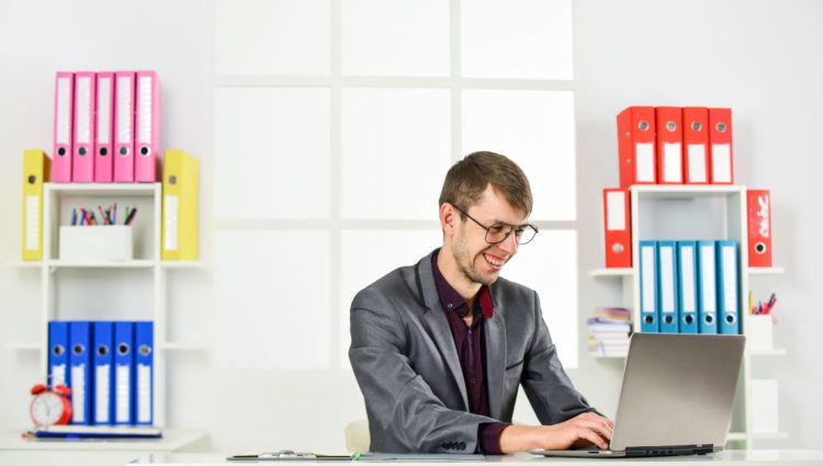 A young man sitting on a desk and working on his laptop, smiling, a pile of folders at the back