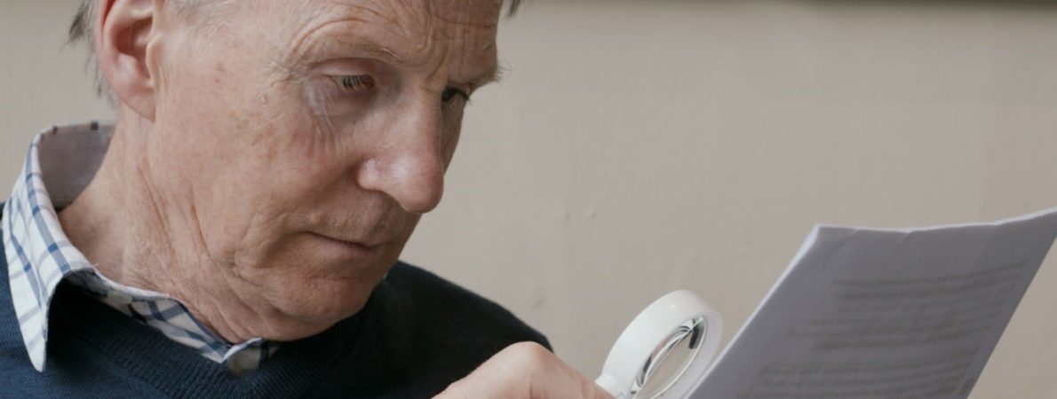 A man in a blue jumper reads a letter using a magnifier