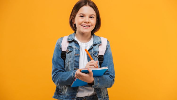 A young girl, holding some books and taking notes while smiling at the camera