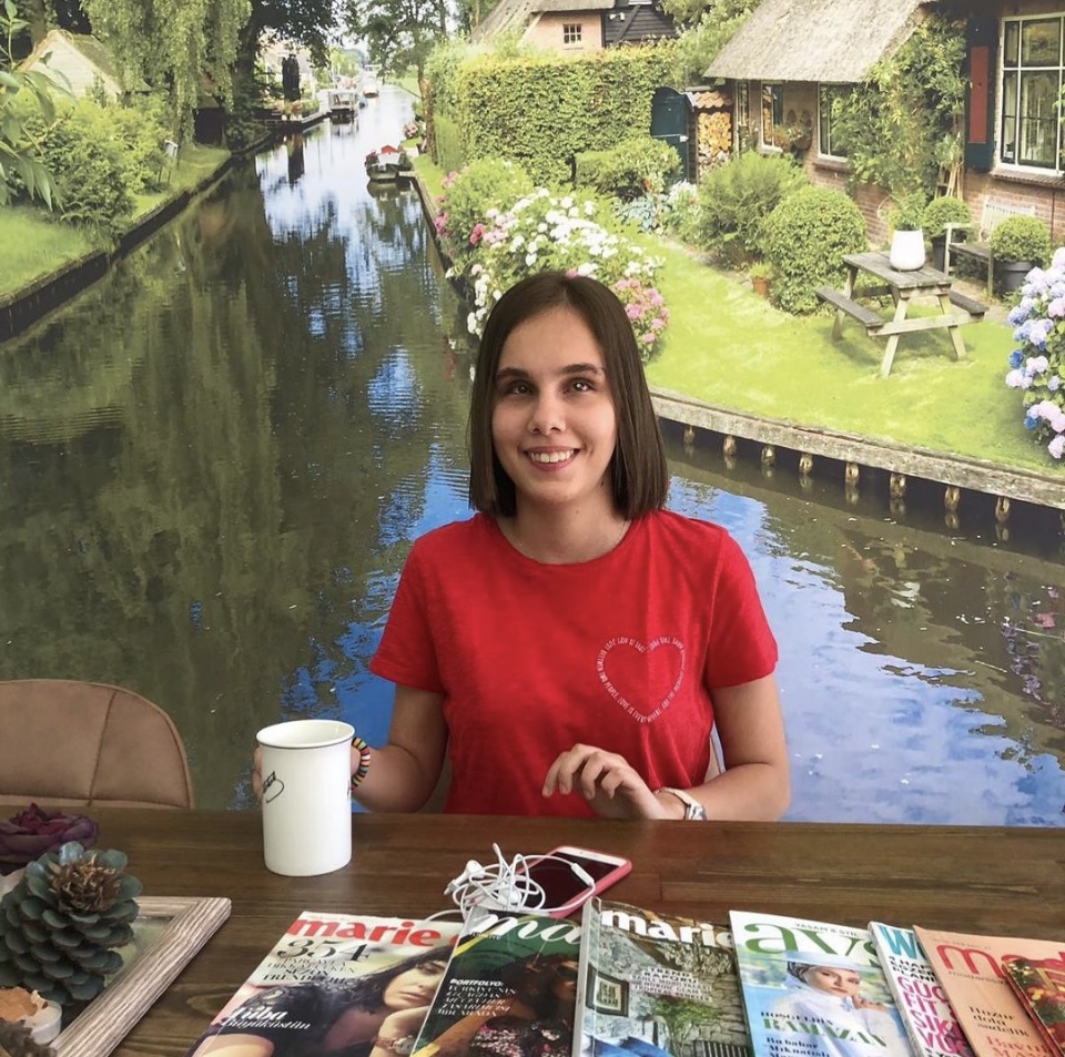 A picture of Zehra sitting on a chair, behind a desk that has some colourful flyers on. She is wearing a red t-shirt, looking at the camera and smiling