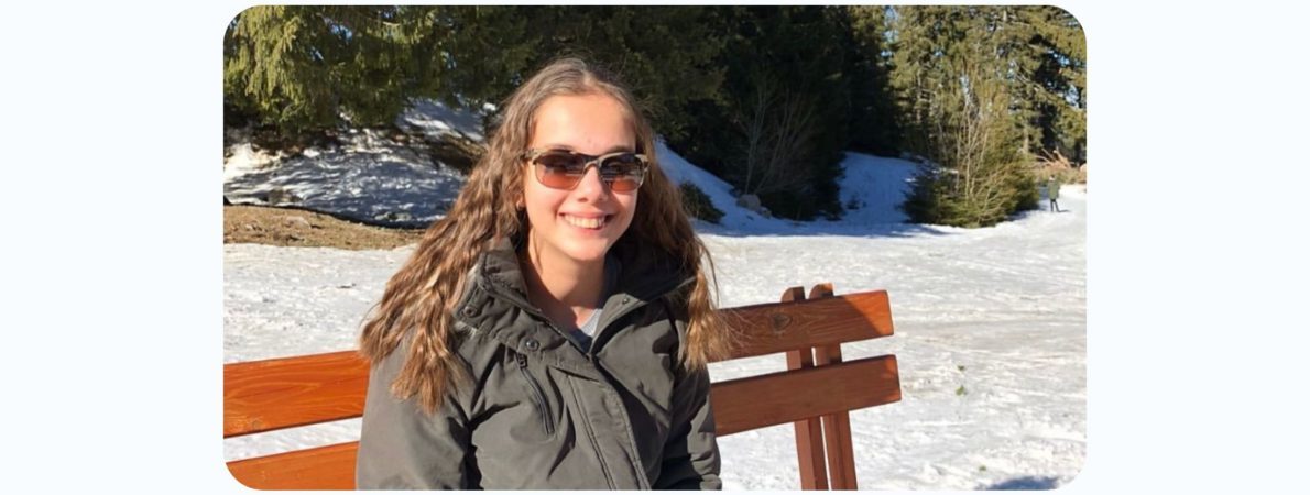 An image of Zehra wearing a winter coat, sitting on a bench, enjoying the snowy background. Looking at the camera and smiling