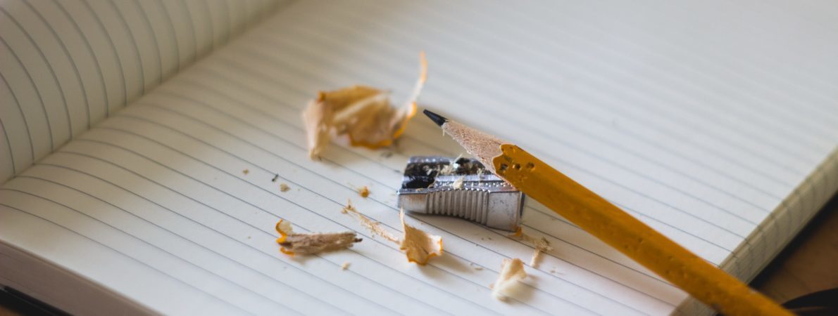 A pencil on an open paper notebook, with a tool for sharpening