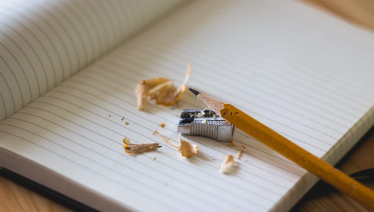 A pencil on an open paper notebook, with a tool for sharpening