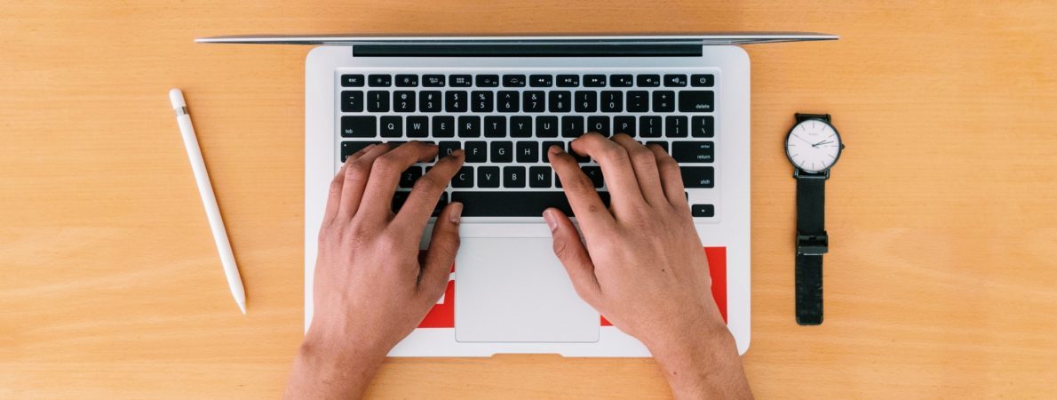 A picture of a person typing on a laptop's keyboard, sat on a wooden desk with a watch on the right and a pencil on the left side.