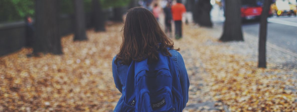 Woman with a blue back bag walking on a street covered on autumn leaves