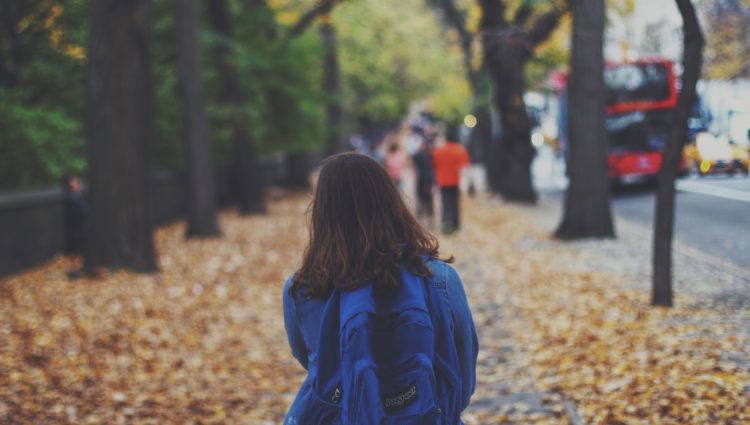 Woman with a blue back bag walking on a street covered on autumn leaves