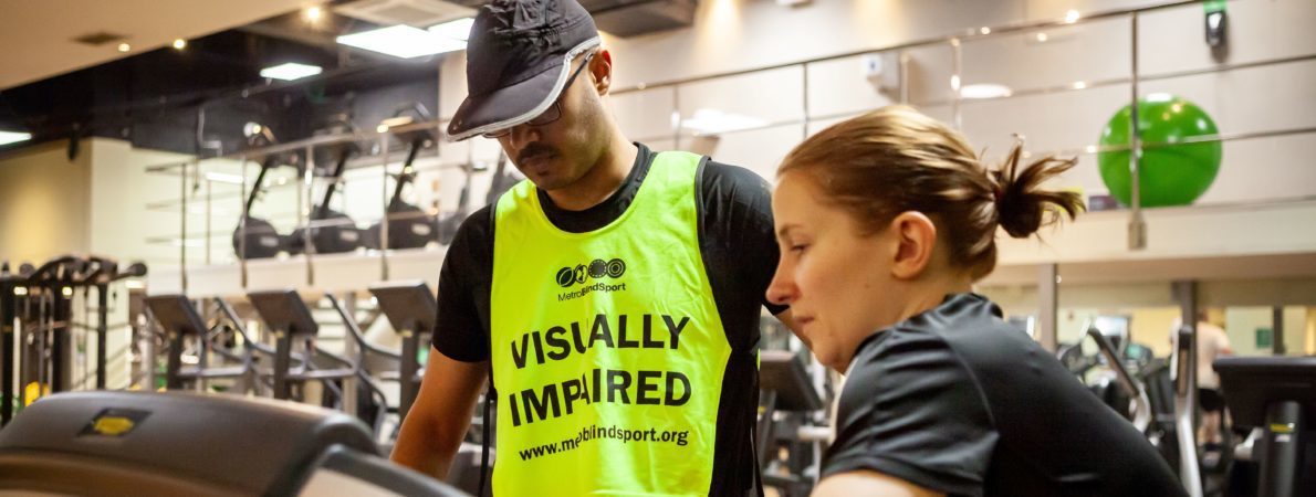 A young man wearing a hat and a vest that says 'Visually impaired' by a treadmill, getting help from a sighted person on how to use it.