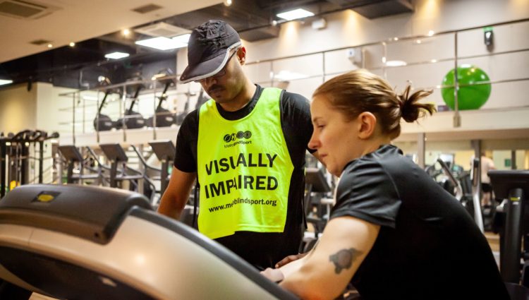 A young man wearing a hat and a vest that says 'Visually impaired' by a treadmill, getting help from a sighted person on how to use it.