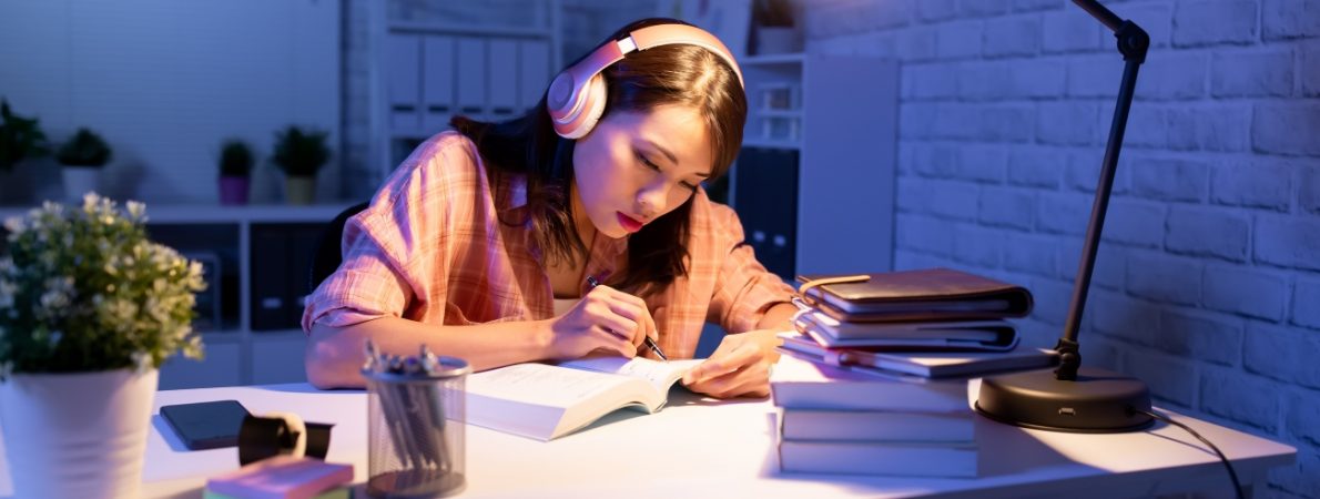 An Asian young woman studying in the evening on her desk with lots of books around her and a lamp on