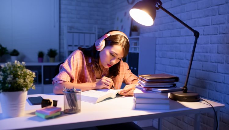 An Asian young woman studying in the evening on her desk with lots of books around her and a lamp on | links to article: Academic Pressure and Stress at University
