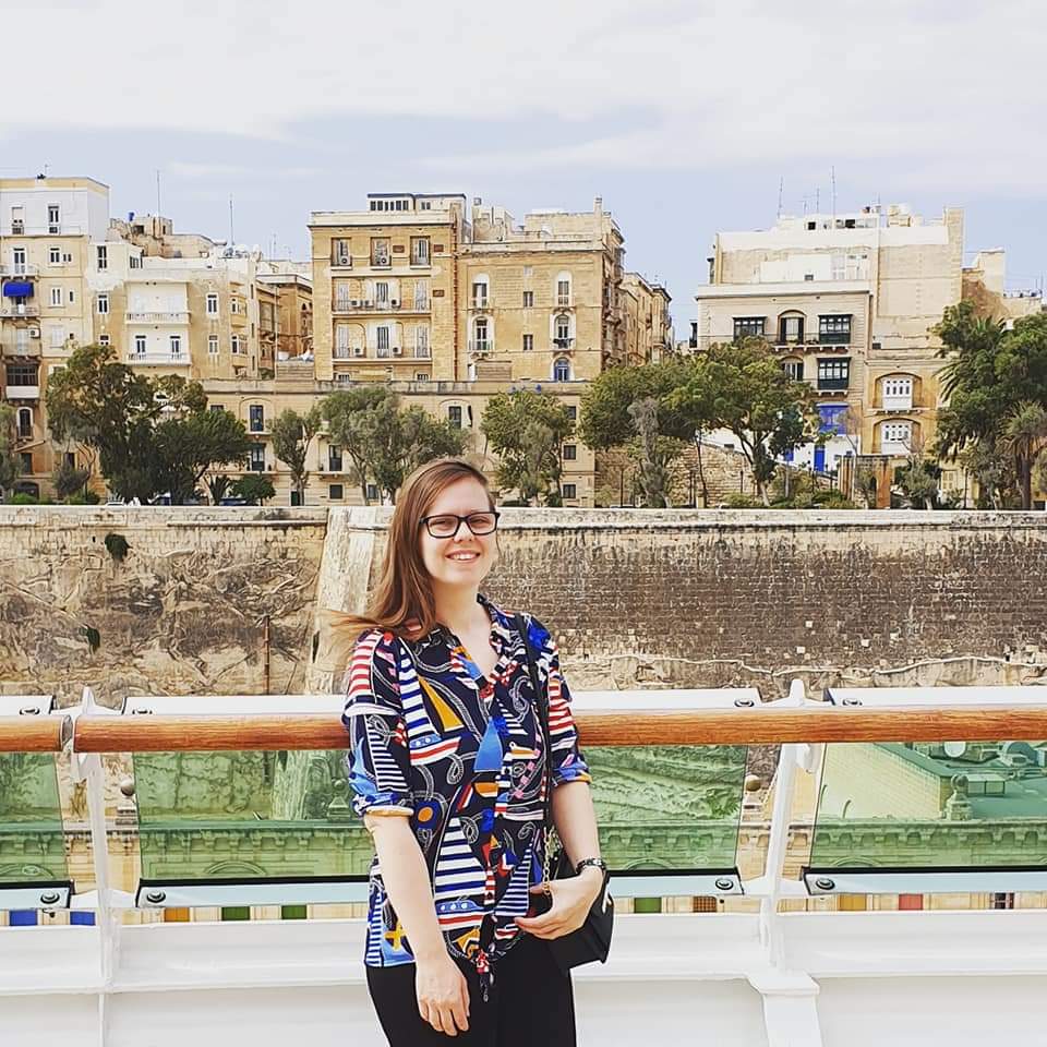 A picture of Fiona standing, looking at the camera and smiling, in front of some buildings during her holidays