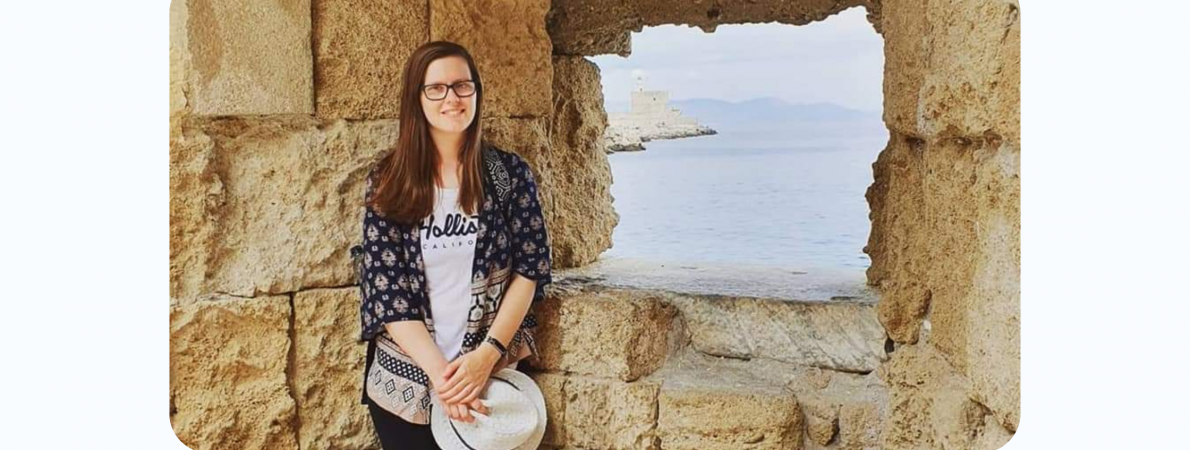 A picture of Fiona standing by a window of an ancient building, most probably a castle. She is looking at the camera and smiling, holding a hat in her hand.