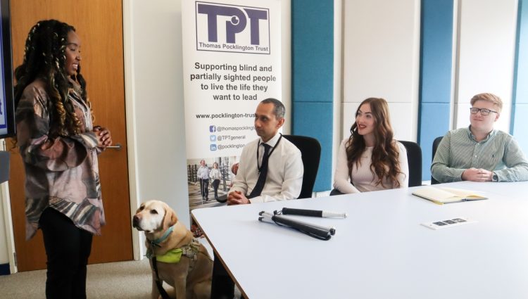 A girl standing and delivering a presentation to three people sitting around a table. A guide dog is sitting on the floor next to her.