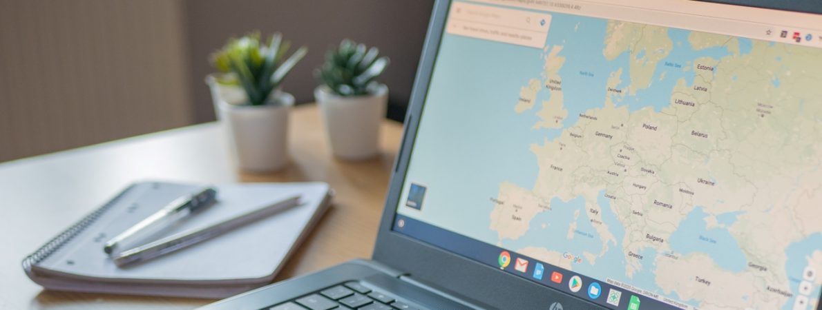 A google chromebook on desk. On the screen there is the map of Europe. A notepad and pens and two small plants next to it on the table