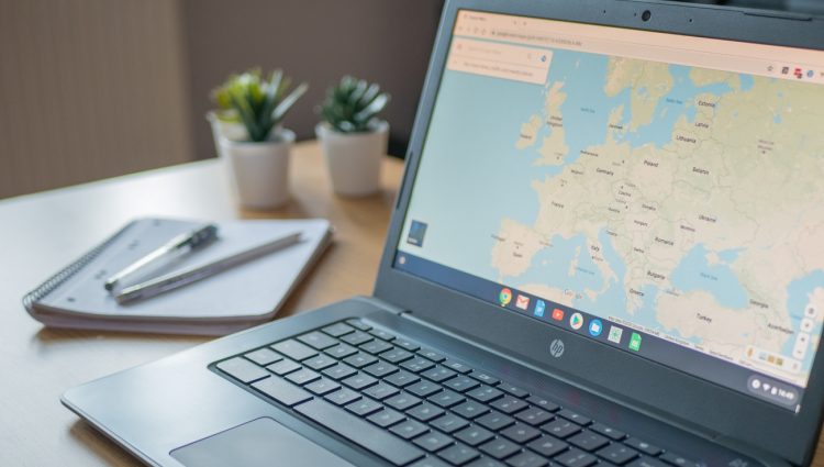 A google chromebook on desk. On the screen there is the map of Europe. A notepad and pens and two small plants next to it on the table
