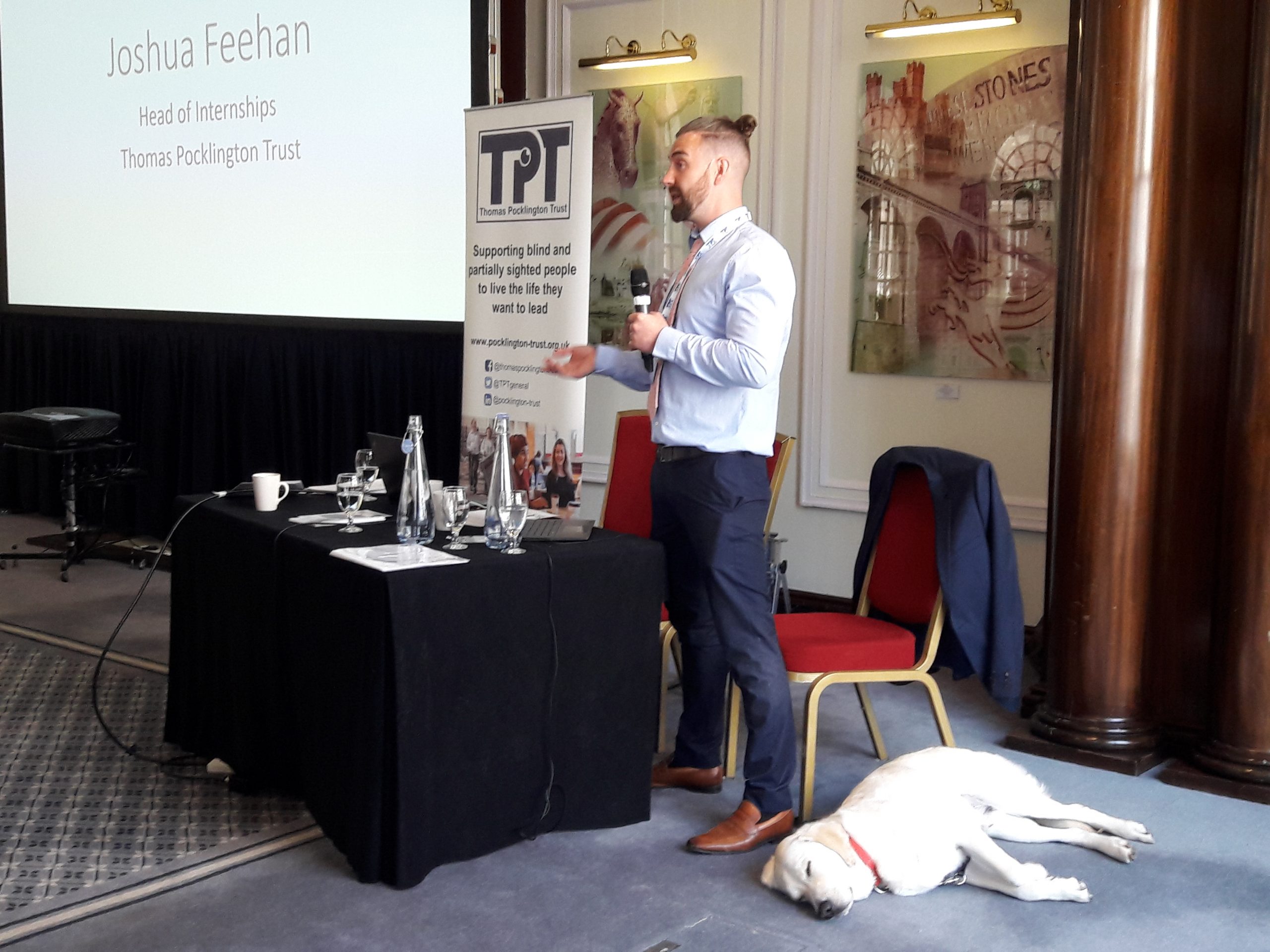 Josh Feehan, TPT's Head of internships standing next to a table and presenting the programme during the event. Next to him, his guide dig Ringo is lying on the floor.