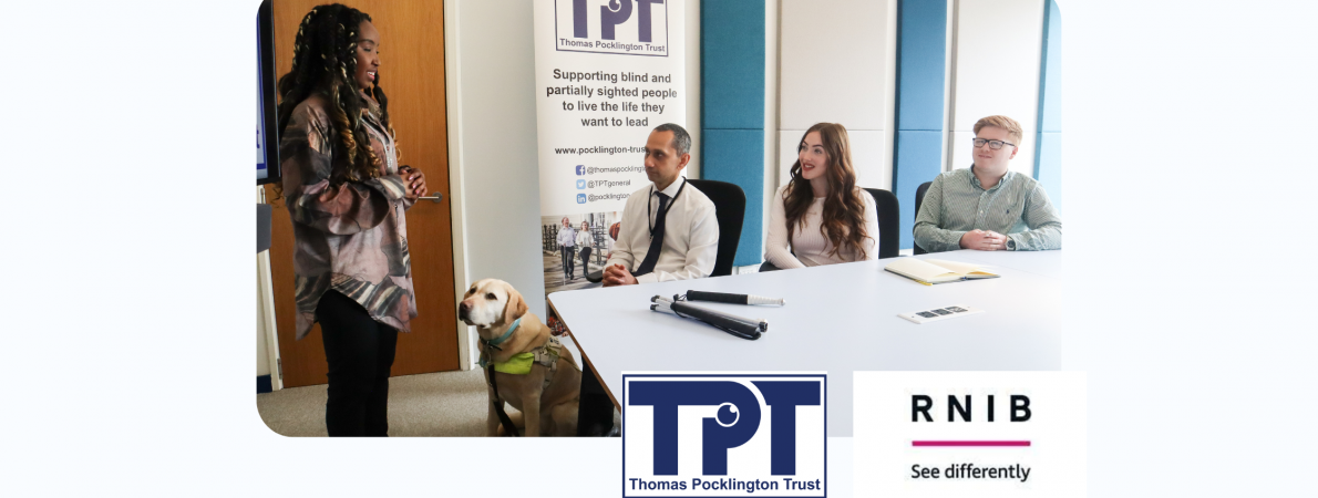 A picture of two young men and a woman sitting around a table in a meeting room. They are all listening to a young lady standing and delivering a presentation. Next to her, there is a guide dog standing. TPT's banner is behind them. TPT and RNIB logos on bottom right
