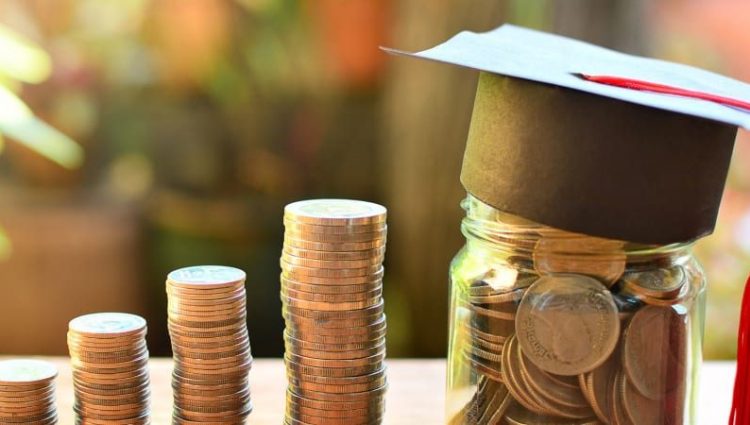 Image with a stack of coins on a table and a jar with savings in it. On top of the jar there is a small graduation hat.