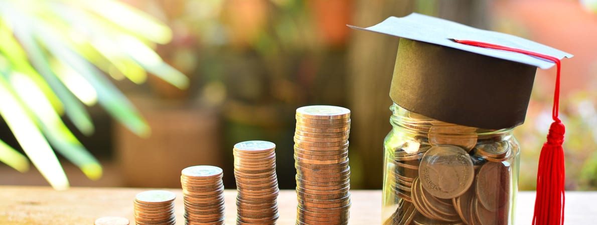 Image with a stack of coins on a table and a jar with savings in it. On top of the jar there is a small graduation hat.