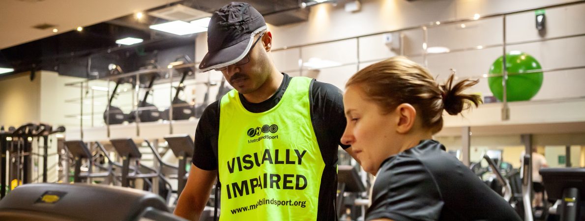 A woman showing a man with a vest that says 'Visually impaired' how to use the treadmill in a gym.